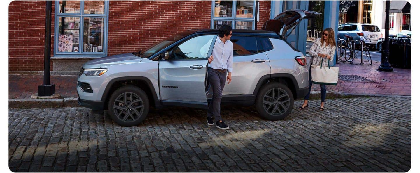 Un Jeep Compass Limited Altitude plateado del año 2026 está aparcado en una calle adoquinada en un distrito comercial, con un hombre saliendo por la puerta del conductor y una mujer de pie junto al portón trasero abierto.
