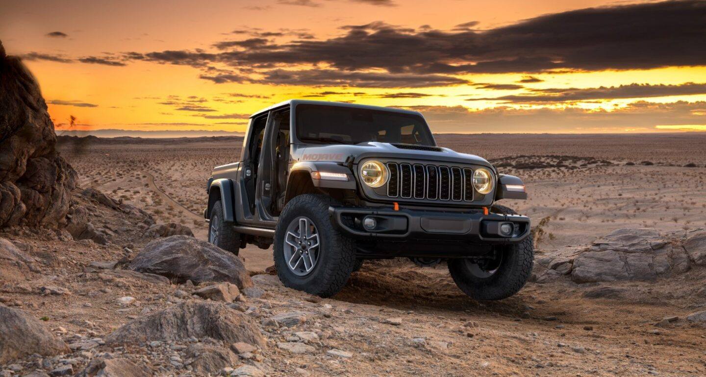Un Jeep Gladiator Mojave X negro 2026 con puertas de tubo, subiendo por un sendero todoterreno en el desierto al atardecer.