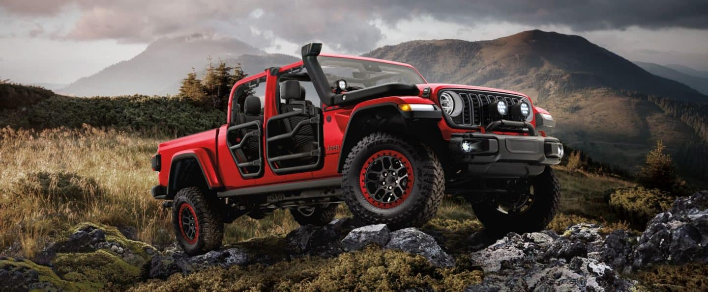 Un Jeep Gladiator Rubicon X 2026 gris con las puertas y el techo extraídos y los focos delanteros y antiniebla encendidos, estacionada en la cima de una colina rocosa. 