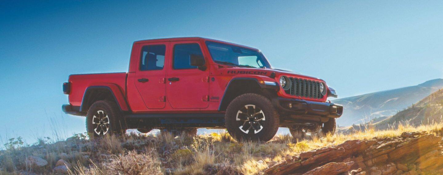 Perfil del lado del pasajero de una Jeep Gladiator Mojave X 2026 gris estacionada en terreno arenoso al atardecer. Sobre la imagen, hay un esténcil de la parrilla de siete ranuras de la marca Jeep. Gladiator. 