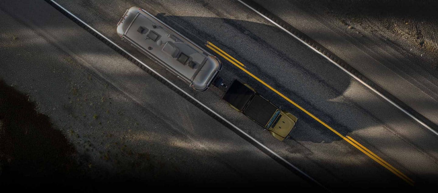 Ángulo aéreo de una Jeep Gladiator 2026 en verde oliva circulando por una carretera, acarreando un gran remolque de viaje. 