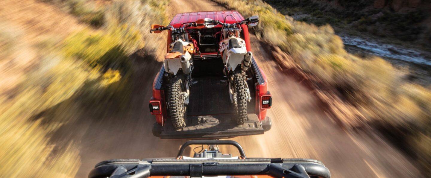 Vista trasera elevada de un Jeep Gladiator Rubicon X rojo del año 2026 circulando por un camino de tierra con curvas, arrastrando un remolque. El portón trasero de la camioneta está abierto con dos motos de cross amarradas en la caja. El fondo está difuminado para indicar la velocidad. 