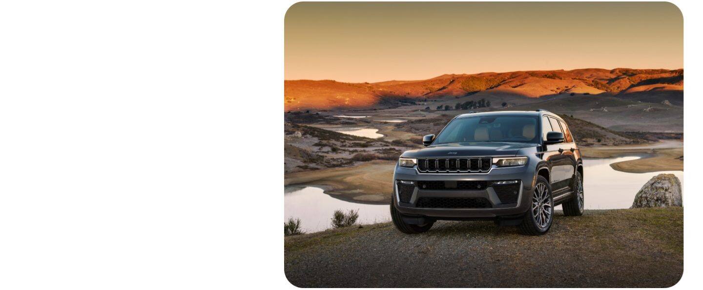 Un Jeep Grand Cherokee Trailhawk 4xe 2026 blanco con una inserción negra en el capó, ingresando a un arroyo desde un sendero cubierto de césped, fuera de la carretera. 