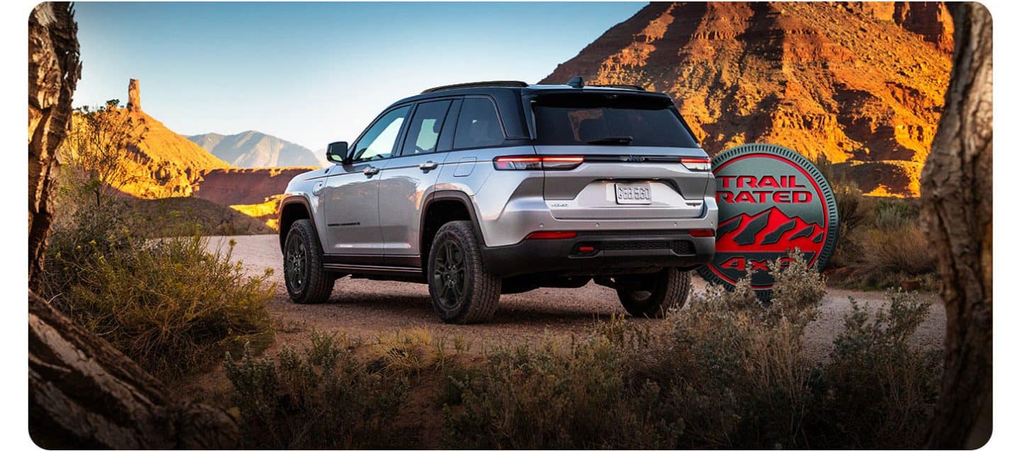 Un ángulo trasero del lado del conductor de un Jeep Grand Cherokee Trailhawk 4xe 2026 plateado estacionado en un claro en el desierto, rodeado de montañas. El emblema Trail Rated 4x4. 