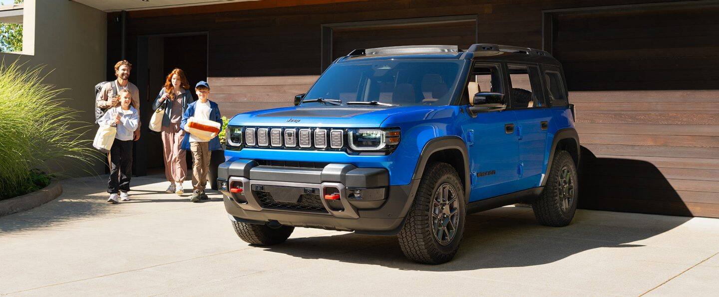 Un Jeep Recon Moab 2026 azul con techo negro, estacionado en la entrada de una casa residencial, con una familia de cuatro personas caminando hacia el vehículo.