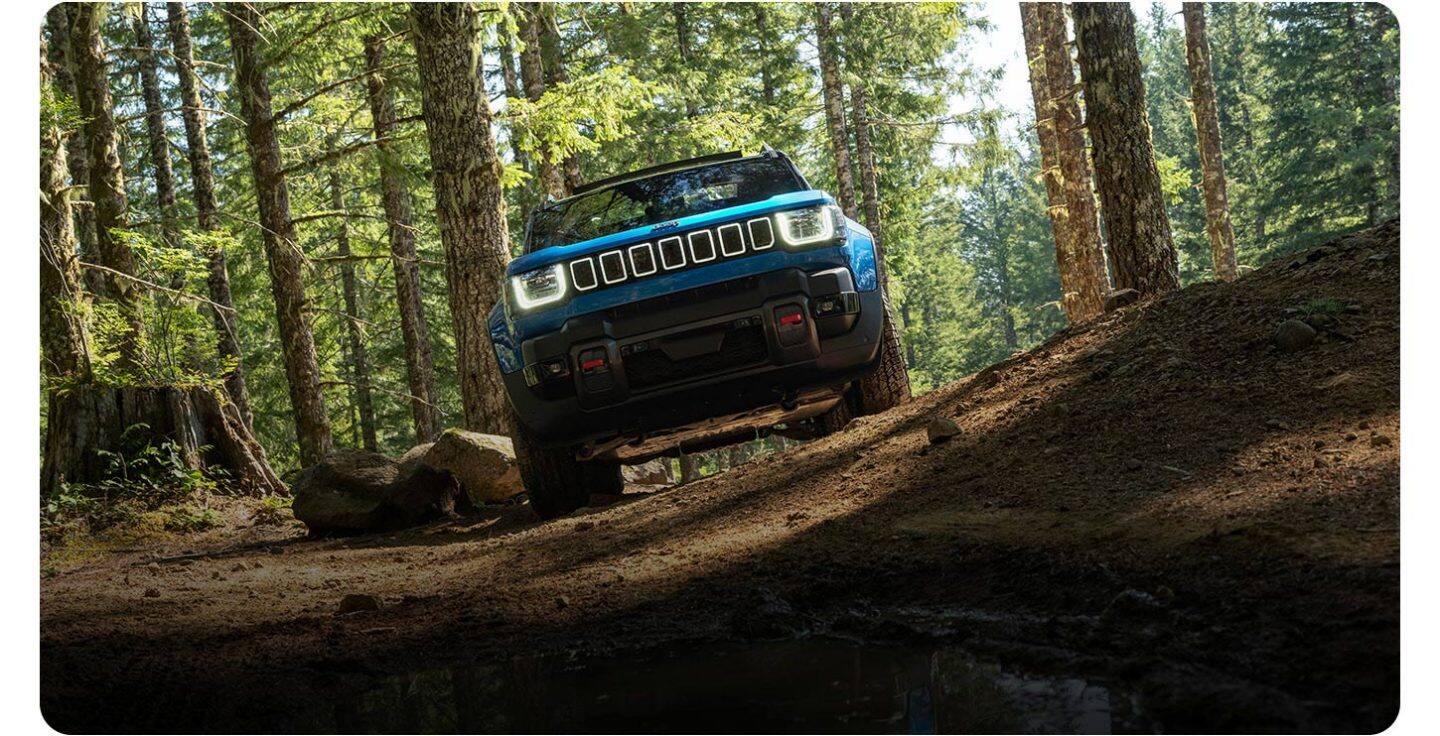 Ángulo frontal bajo de un Jeep Recon Moab 2026 azul viajando horizontalmente a través de una colina en el bosque.