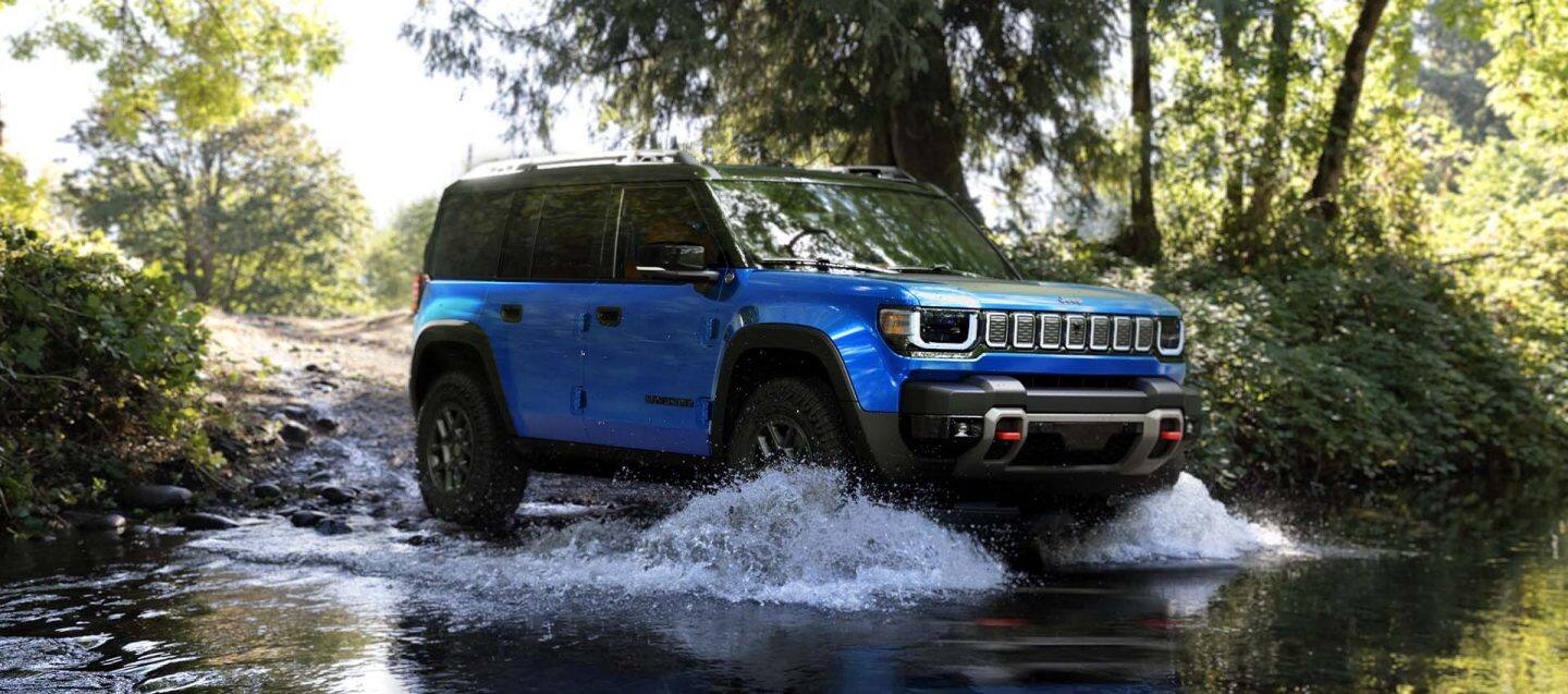 Ángulo frontal del lado del pasajero de un Jeep Recon Moab 2026 azul con techo negro todoterreno, entrando en un arroyo.