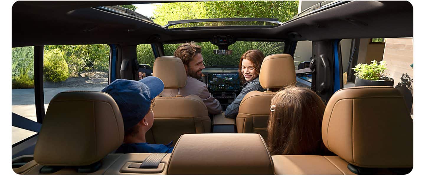 El interior de un Jeep Recon Moab 2026 con el techo corredizo abierto, el papá en el asiento del conductor y la mamá en el asiento del pasajero delantero, ambos mirando hacia atrás al niño y la niña en los asientos traseros. Toma desde la parte trasera del SUV.