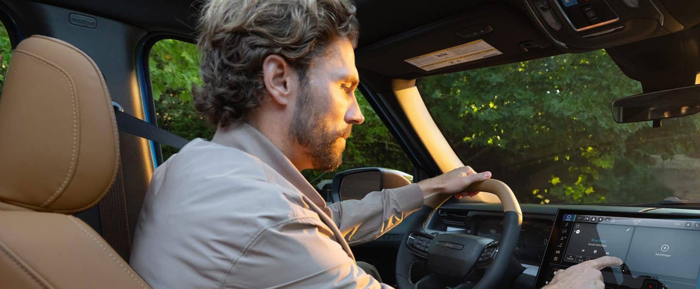 Un hombre en el asiento del conductor de un Jeep Recon Moab 2026 con una mano en el guía y la otra haciendo una selección en la pantalla táctil Uconnect.