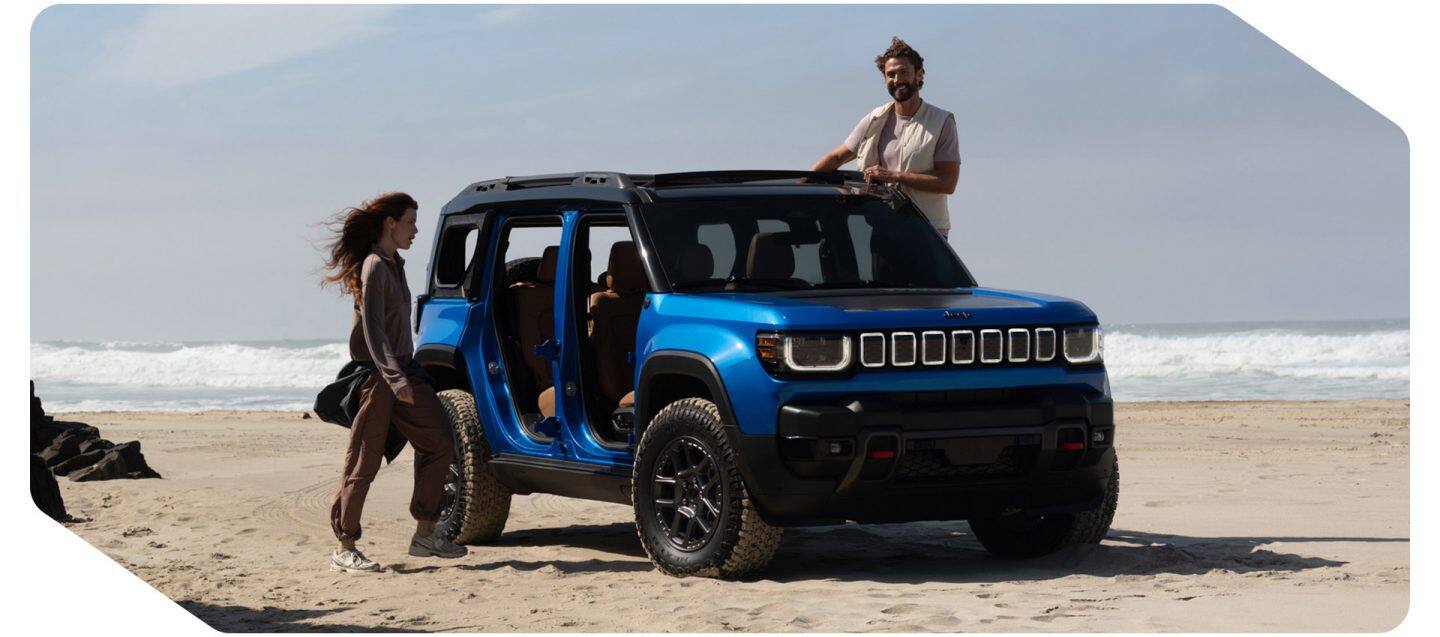 Una toma frontal del lado del pasajero de un Jeep Recon Moab 2026 azul con las puertas removidas, estacionado en una playa de arena con una mujer cerca del lado del pasajero y un hombre de pie en el umbral de la puerta del lado del conductor del SUV.