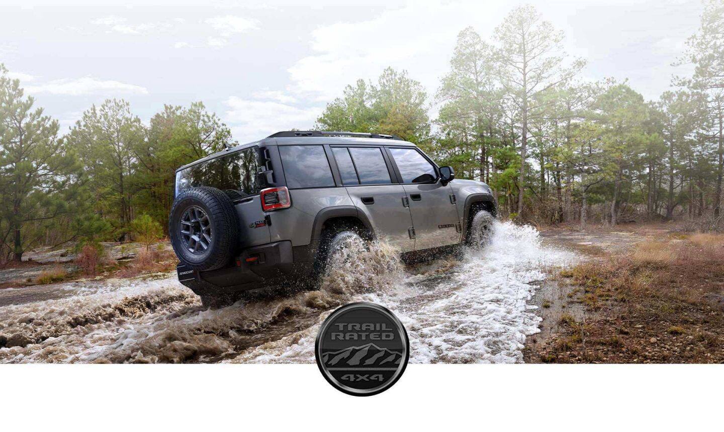 Ángulo trasero del lado del pasajero de un Jeep Recon Moab 2026 gris vadeando un arroyo fangoso. El emblema Trail Rated 4x4.