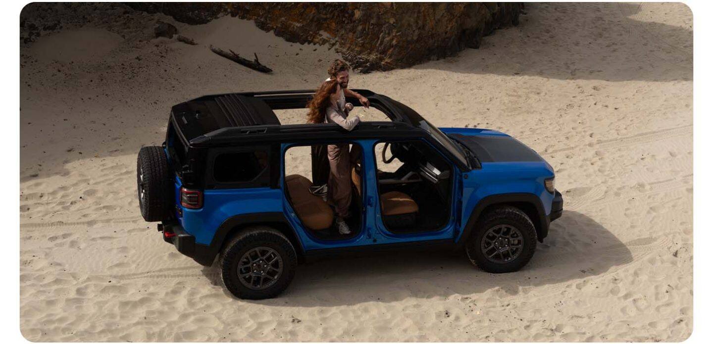 Una vista elevada del lado del pasajero de un Jeep Recon 2026 azul con las puertas removidas, estacionado en una playa de arena. Su techo eléctrico se muestra abierto y una pareja está de pie en el piso de la segunda fila, con la parte superior del cuerpo asomándose por la abertura del techo corredizo. Hay tres puntos interactivos seleccionables en el SUV.