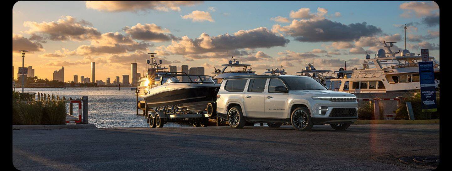 Una Jeep Grand Wagoneer Summit Reserve Obsidian 2026 blanca sacando una lancha motora del agua en una rampa de embarcaciones junto al río al atardecer.