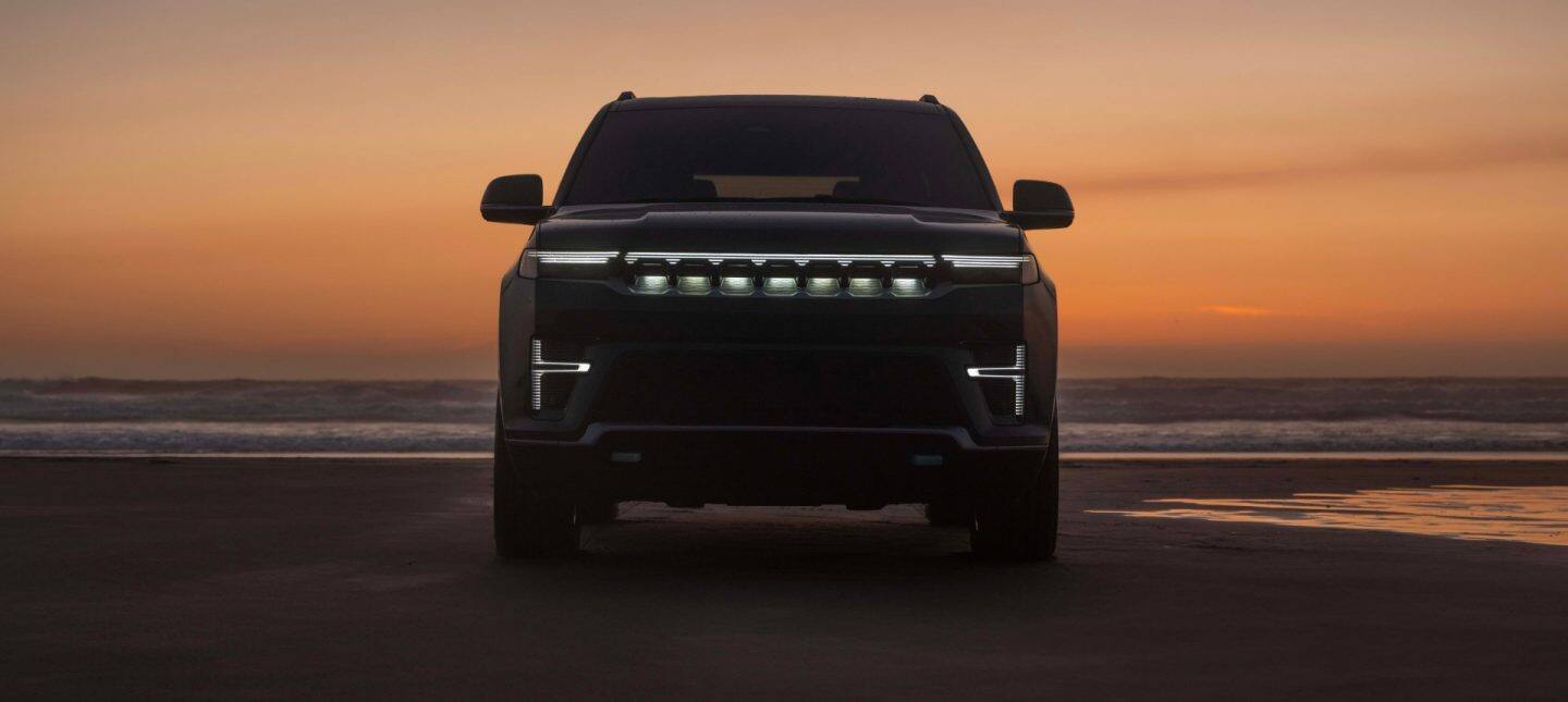 Vista frontal de una Jeep Grand Wagoneer Upland 2026 negra conduciendo por una playa de arena junto a la orilla al atardecer, levantando agua a su paso.