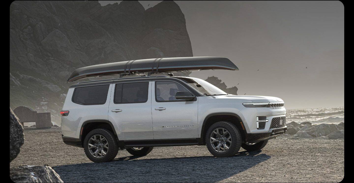 Vista lateral del lado del pasajero de una Jeep Grand Wagoneer Upland 2026 blanca estacionada en una playa rocosa, con un kayak sujeto al portaequipajes del techo.