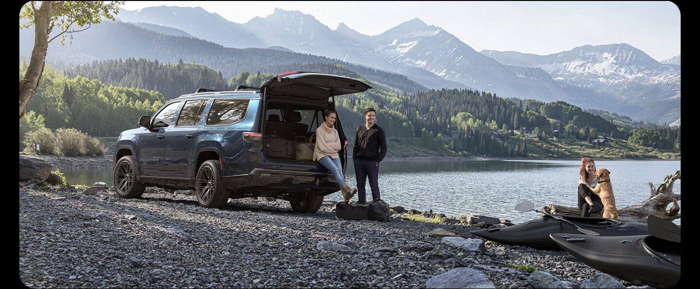 Un Jeep Grand Wagoneer Summit Reserve Obsidian azul de 2026 está aparcado junto a un lago de montaña con la puerta trasera abierta, una mujer está apoyada en el parachoques trasero, un hombre está de pie cerca y una niña con un perro se encuentran a la orilla del agua.
