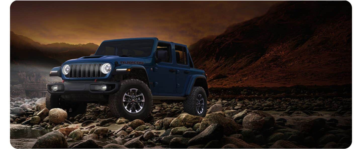 Un Jeep Wrangler Rubicon X 2026 azul de cuatro puertas, con el techo extraído y los faros delanteros encendidos, estacionado en una costa rocosa a la orilla del agua, por la noche.