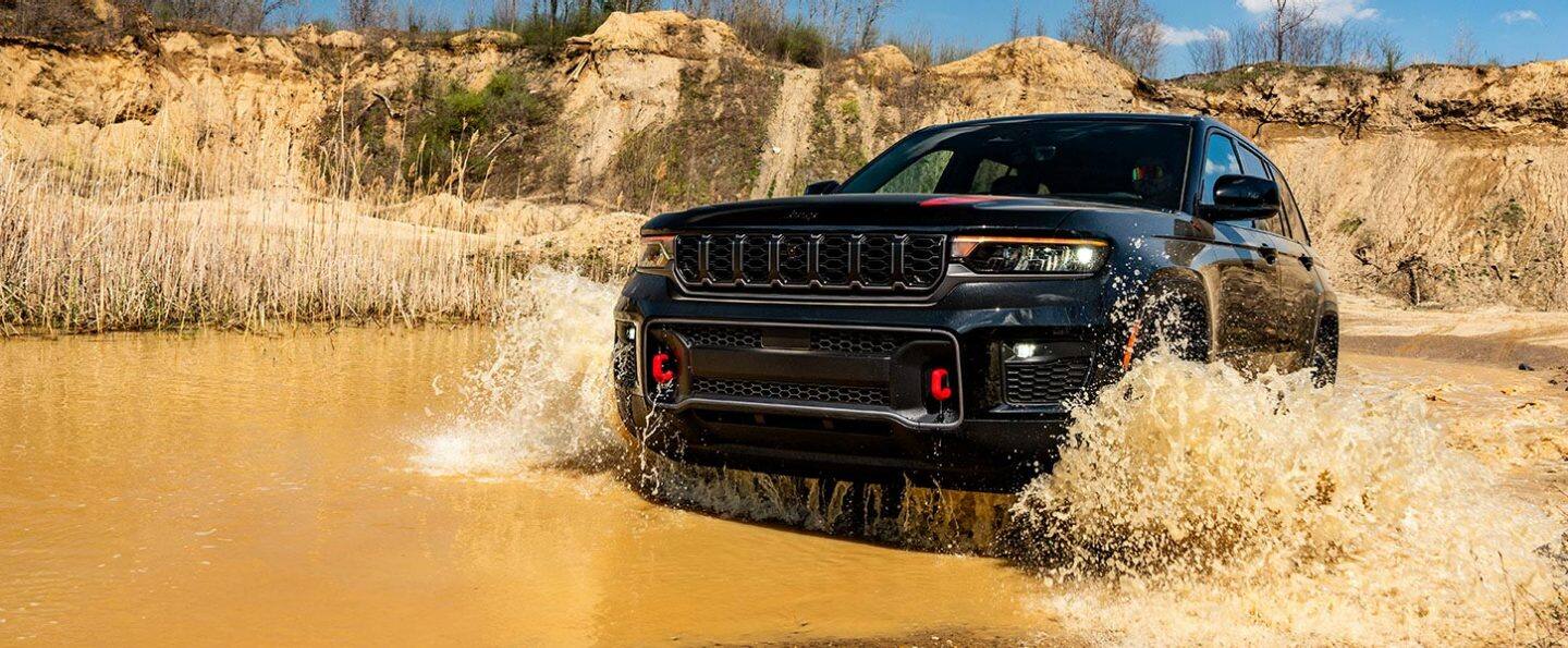 Una Jeep Grand Cherokee Trailhawk 2027 negro vadeando un arroy embarrado, salpicando agua hasta la parte superior de las cavidades para las gomas a medida que avanza.