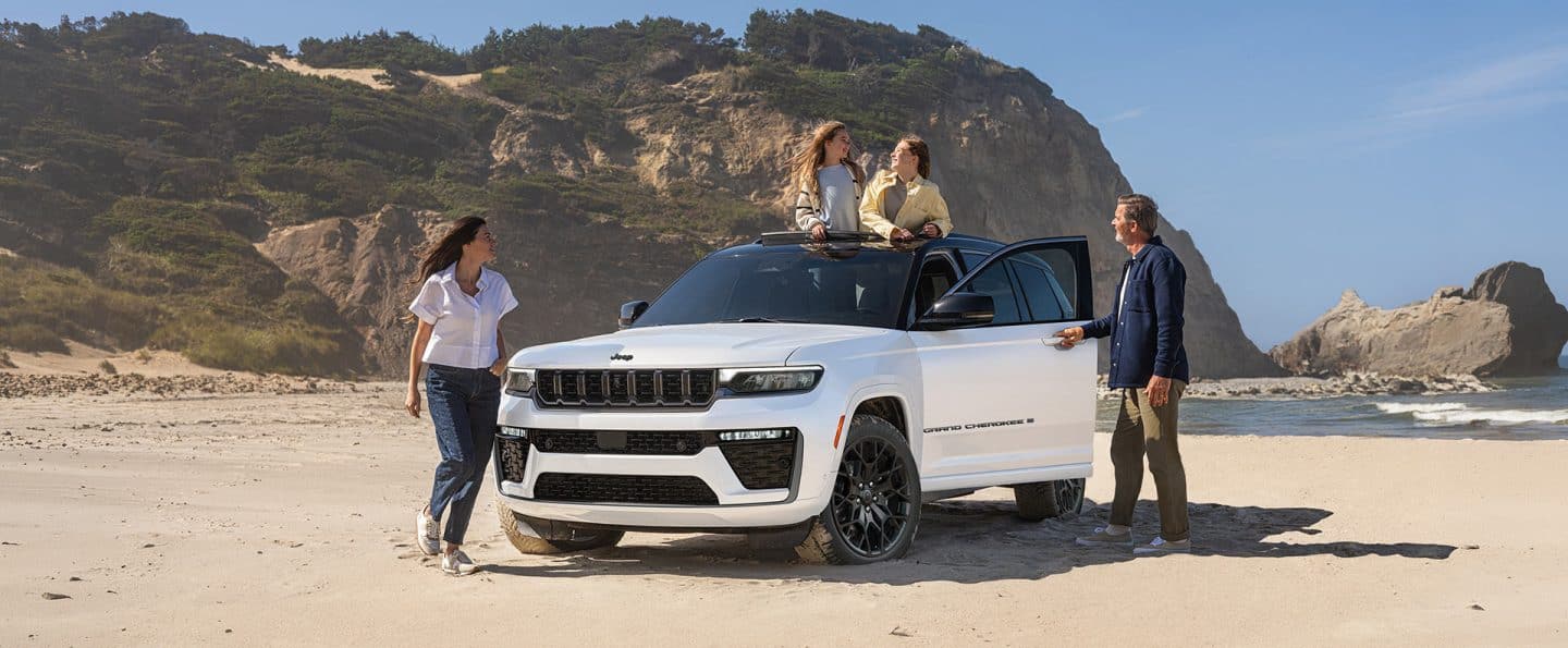 Una Jeep Grand Cherokee Summit blanco está estacionado en la playa, rodeado de colinas cubiertas de césped, mientras una familia sale por las puertas del vehículo y el techo panorámico.