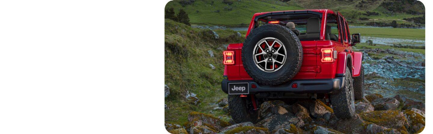 Jeep Wrangler rojo sin techo visto de atrás, escalando rocas en un paisaje montañoso junto a un río.