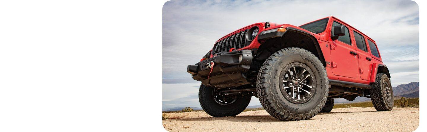 Jeep Wrangler Rubicon rojo con suspensión elevada y llantas todo terreno, vista de tres cuartos delantera en terreno desértico.