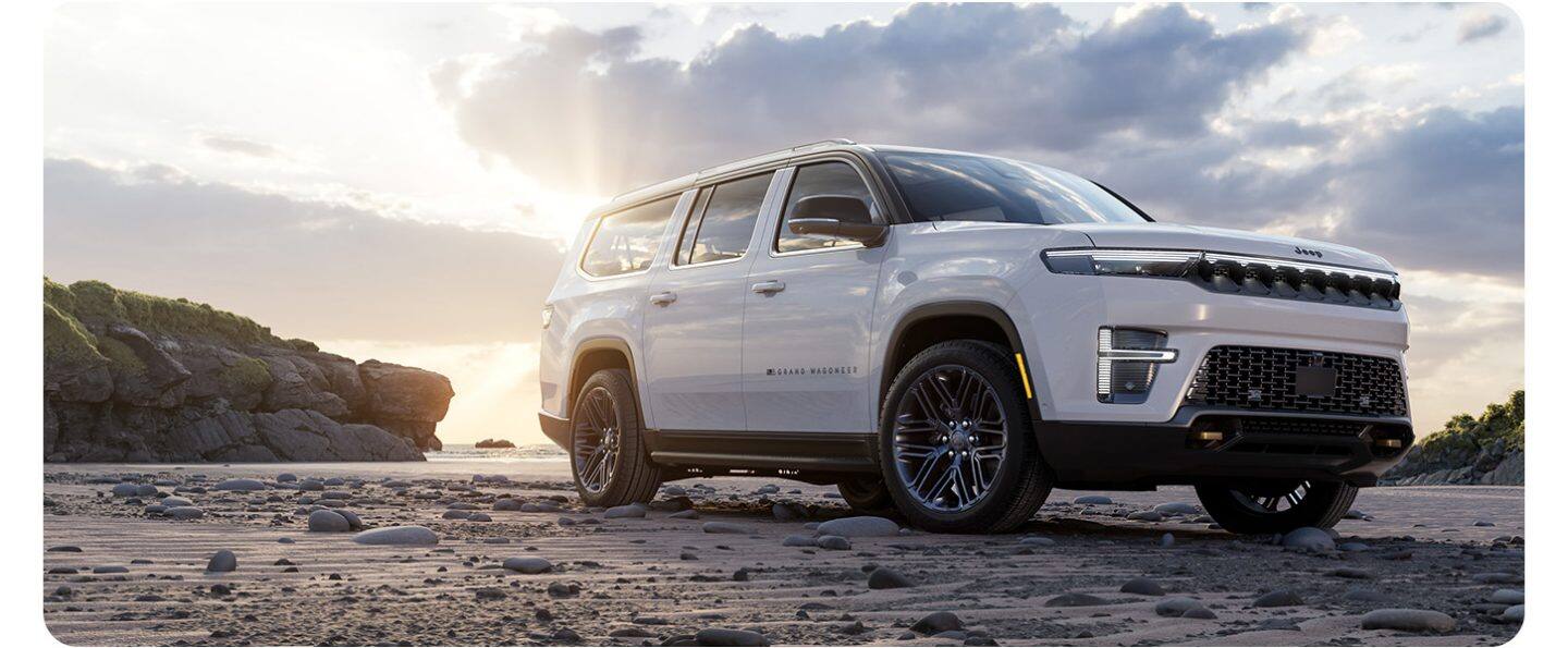 Ángulo frontal inferior del lado del pasajero de un Jeep Grand Wagoneer 2026 Edición 85.° Aniversario blanco estacionado en una playa rocosa al atardecer.