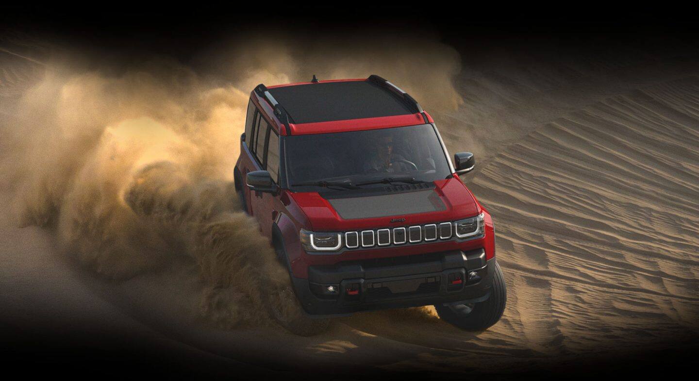 Una Jeep Recon Moab  2026 de color rojo con techo retráctil eléctrico negro y una inserción negra en el bonete, atravesando una duna, con nubes de arena desprendiéndose de sus gomas.