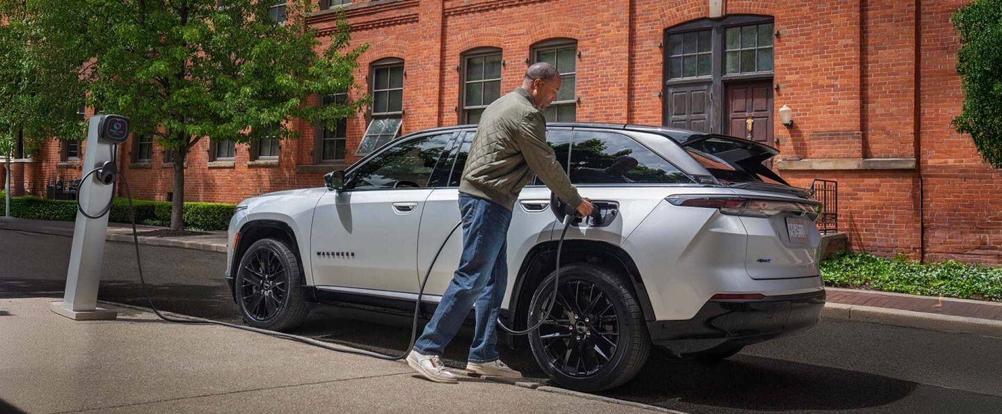 Una Jeep Wagoneer S Launch Edition 2024 plateado estacionado al lado de un edificio de ladrillo, con un hombre enchufando el cable de carga de una estación de carga pública en el puerto de carga del vehículo.