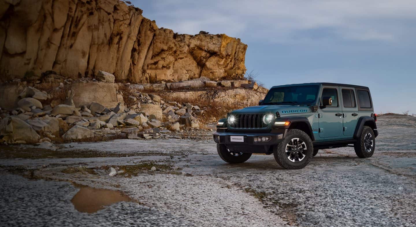 2024 Jeep Wrangler Rubicon 4xe, mille uksed ja katus on eemaldatud, sõidetakse liivarajal maastikul, mõlemal küljel on suured rahnud.