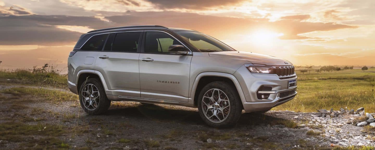 The Trail Ratted 4x4 badge. A blue 2023 Jeep Cherokee Trailhawk kicking up sand on a beach beside crystal blue water.