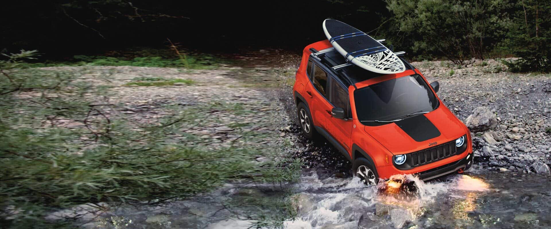 The 2021 Jeep Renegade fording a shallow stream with a surfboard attached to its roof rack.