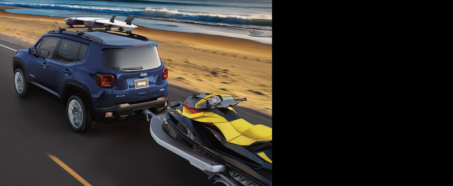 2021 Jeep Renegade tows an ATV and carries a surfboard on its roof rack as it is driven on a beachside road.