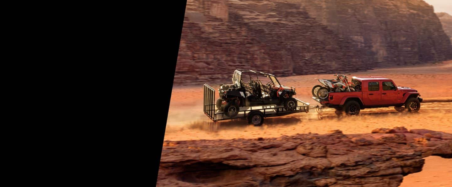 La Jeep Gladiator Rubicon 2021 andando todoterreno con dos motos de cross en la plataforma y remolcando un tráiler con un UTV adentro.