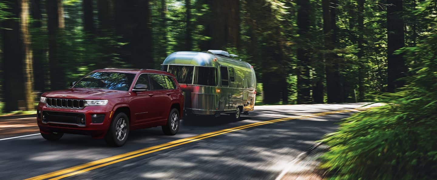 The 2021 Jeep Grand Cherokee L Overland towing an Airstream-style trailer on a two-lane road in the woods.