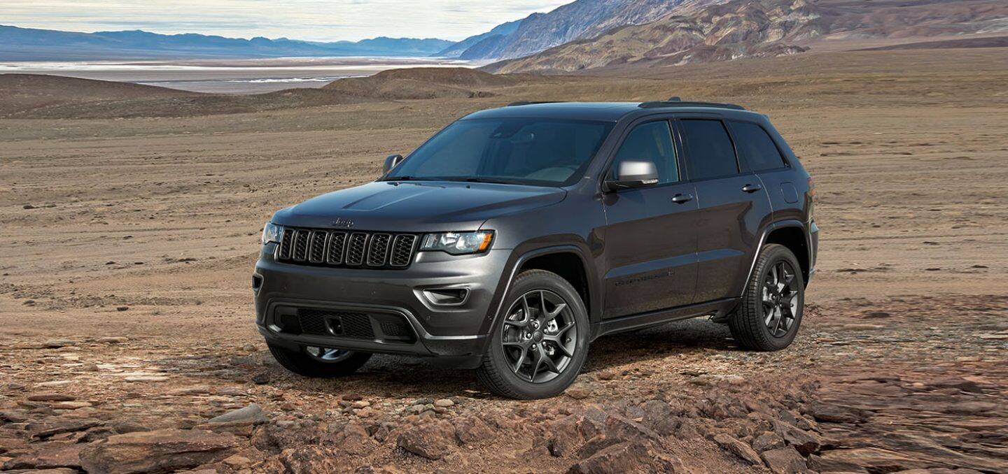 Display A three-quarter front view of a 2021 Jeep Grand Cherokee 80th Anniversary Edition parked on sand with mountains and a body off water in the distance.