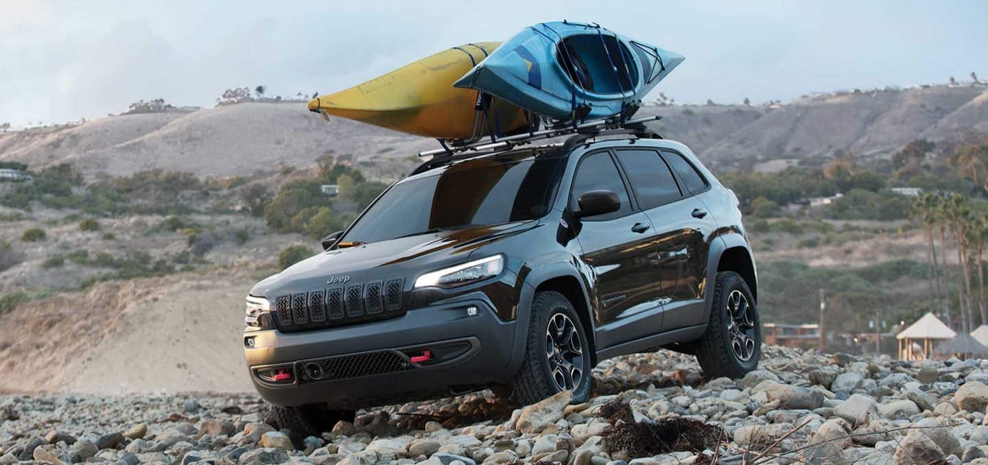 Display The 2022 Jeep Cherokee Trailhawk being driven on a pebbled beach with two kayaks strapped to its roof rack.