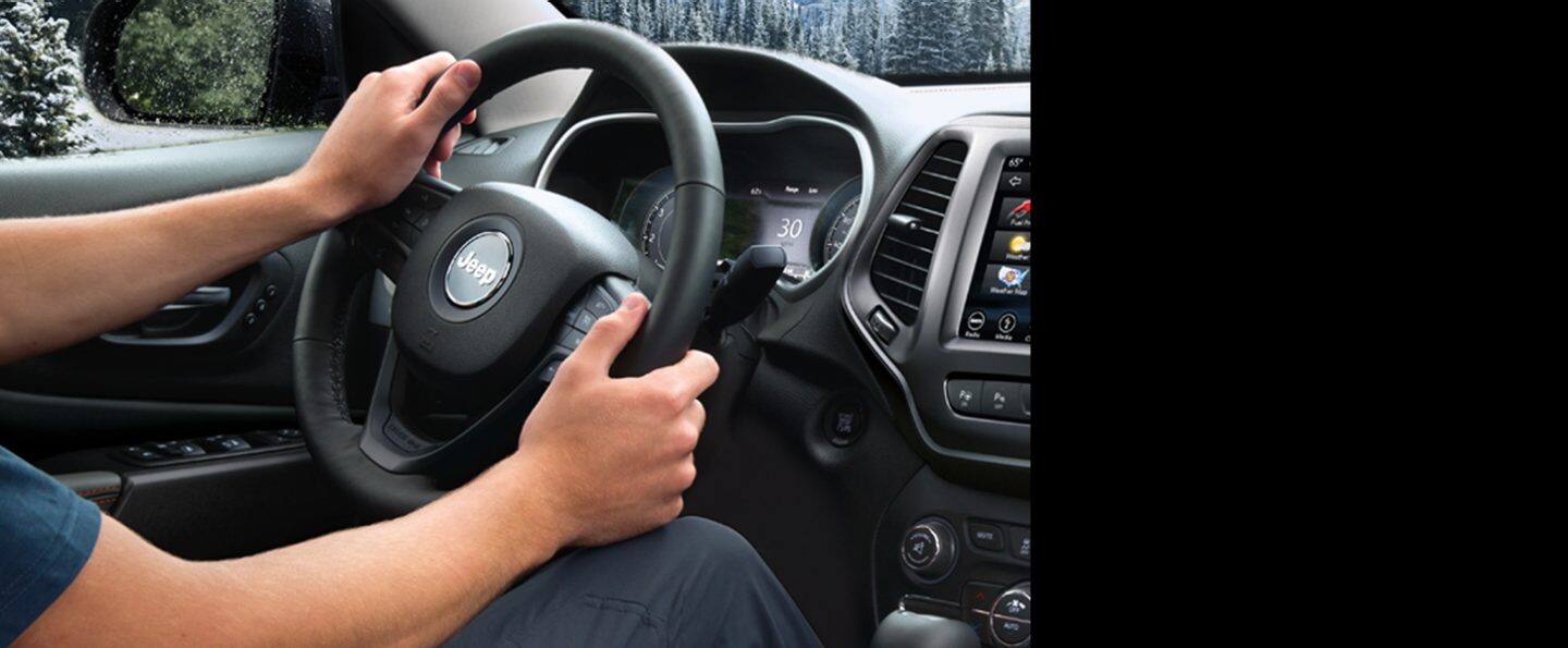 A T-shirt-clad driver grips the wheel in a 2022 Jeep Cherokee while driving through snow-covered woods.