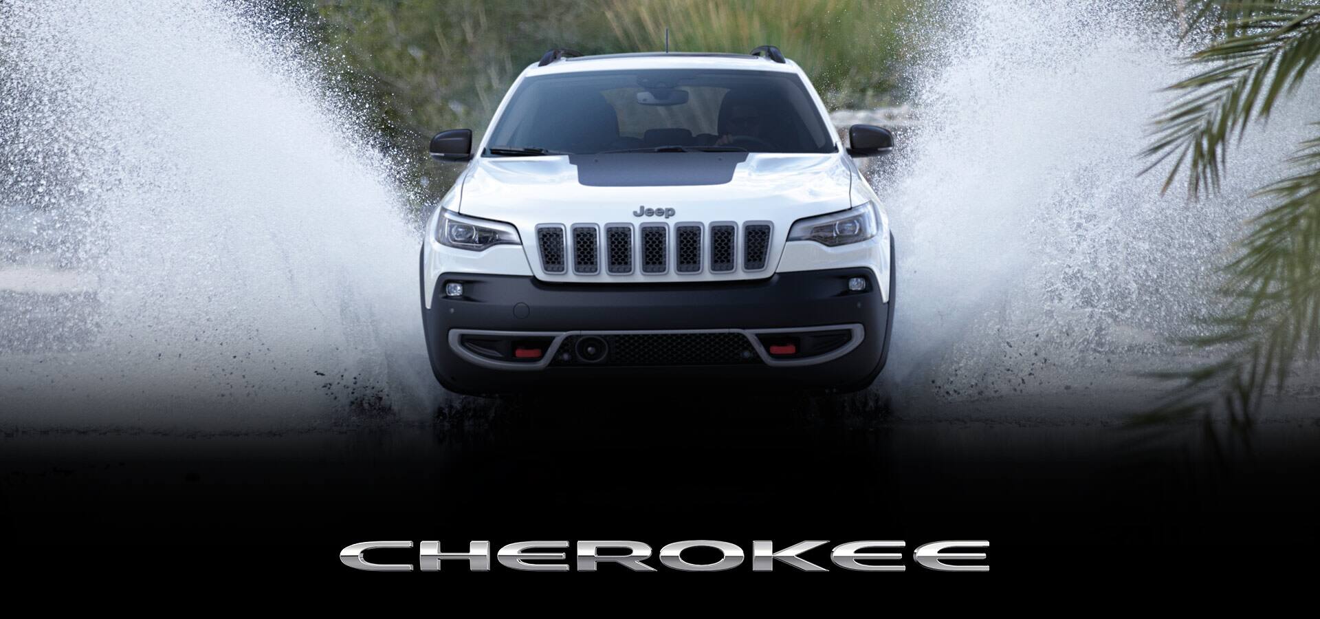 A head-on view of a white 2022 Jeep Cherokee Trailhawk fording a stream, spraying water beyond the height of the vehicle. Cherokee.
