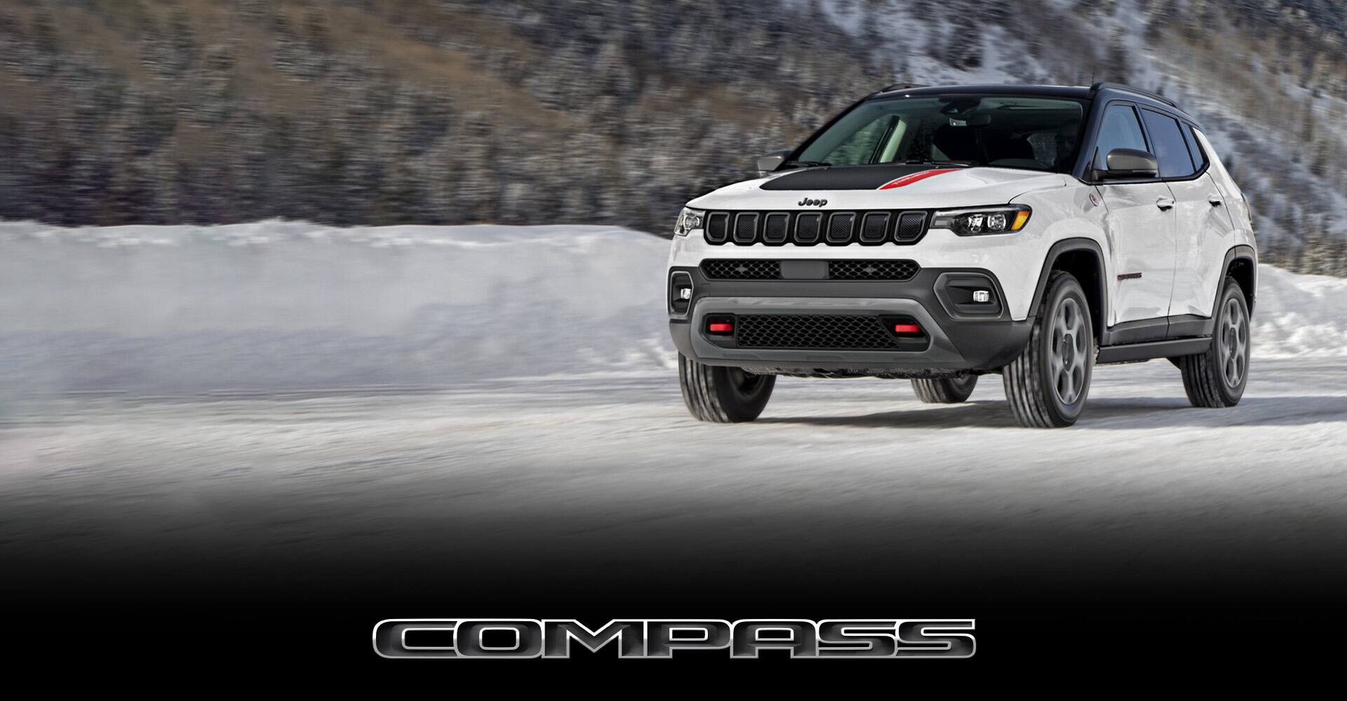 An angled profile of a 2022 Jeep Compass Trailhawk being driven on a snow-covered road beside a mountainous forest that is blurred to indicate the vehicle is in motion. Compass.