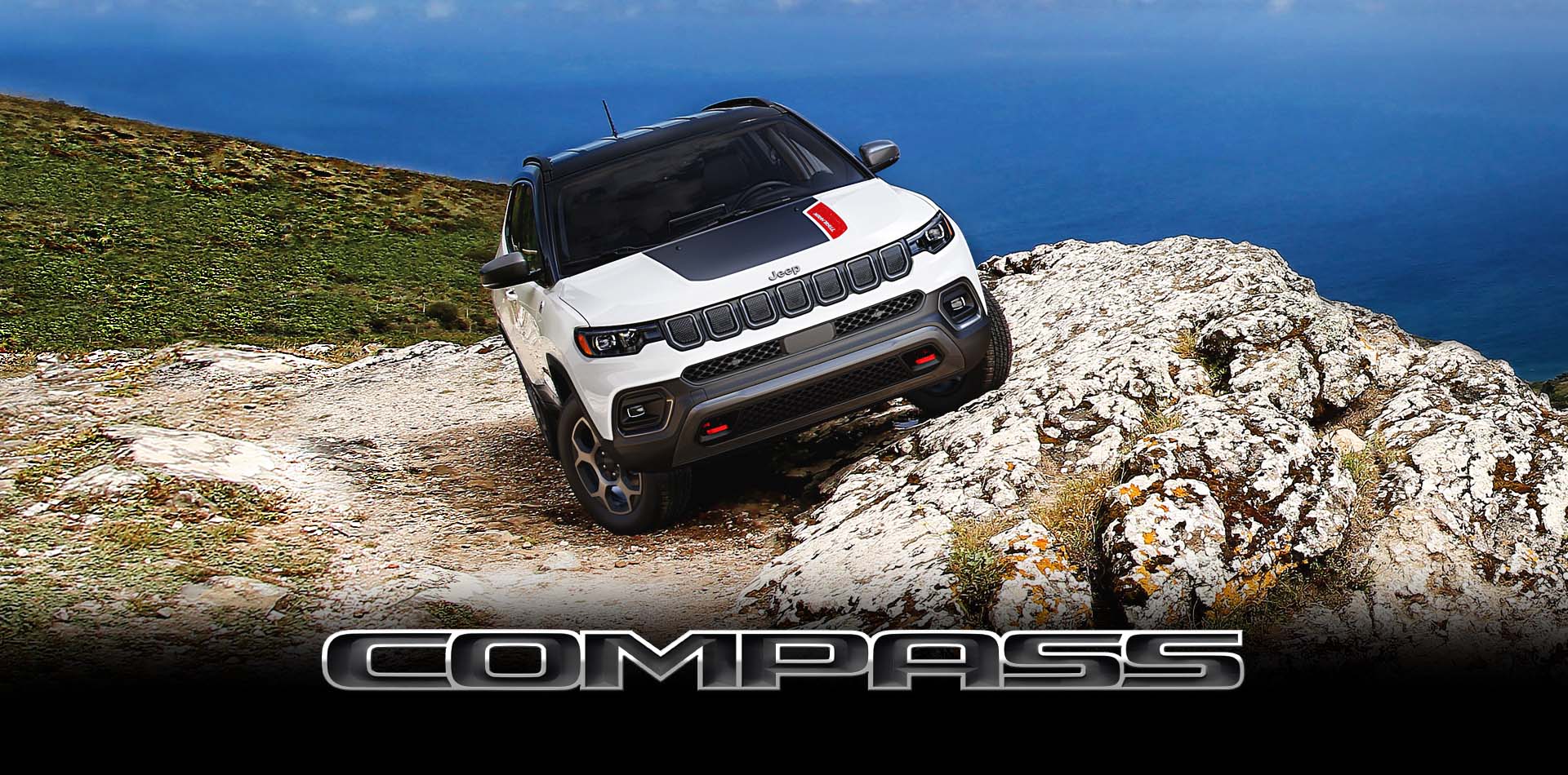 A white 2022 Jeep Compass Trailhawk parked on the slanted edge of an oceanside cliff, its driver-side angled higher than its passenger side. Compass. 