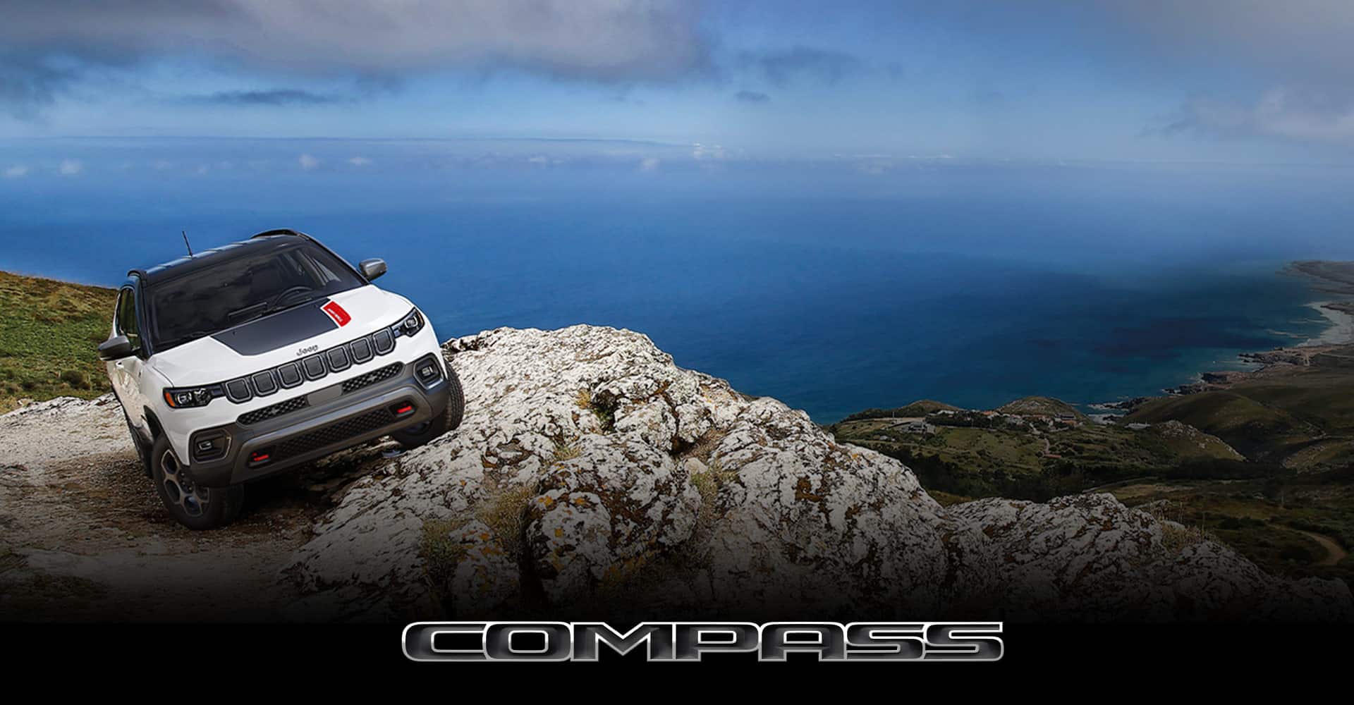 A white 2022 Jeep Compass Trailhawk parked on the slanted edge of an oceanside cliff, its driver-side angled higher than its passenger side. Compass.