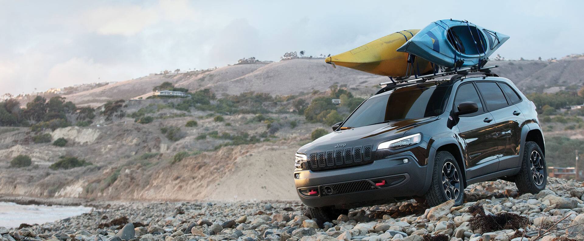 The 2022 Jeep Cherokee being driven on a rocky beach with two kayaks attached to its roof rack.