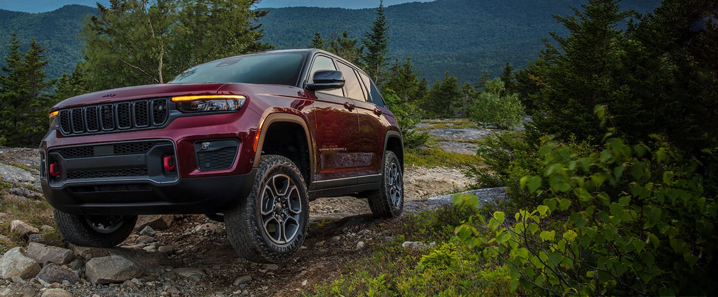 A 2022 Jeep Grand Cherokee Trailhawk parked on a rocky incline at an angle that highlights the vehicle's available ground clearance. Forested hills are visible in the background.