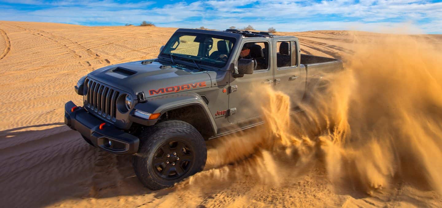 Display The 2022 Jeep Gladiator Mojave being driven on a sand dune, clouds of sand obscuring its rear wheel.