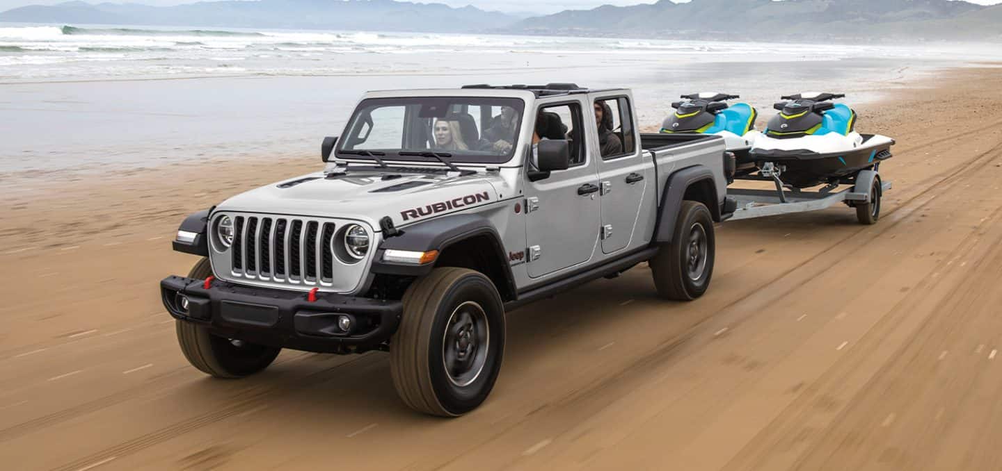 Display A 2022 Jeep Gladiator Rubicon towing two jet skis as it is driven along a beach near the water.