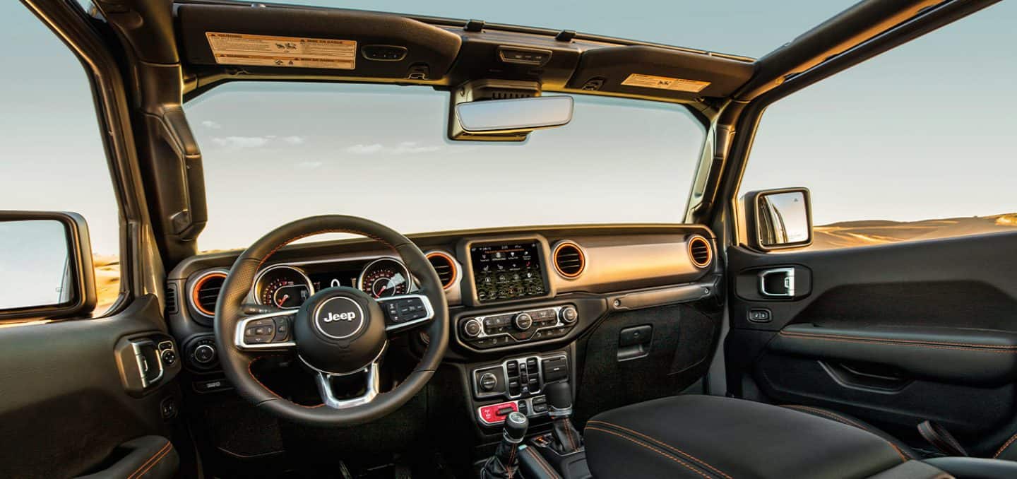 The view from the front row of the 2022 Jeep Gladiator Mojave, focusing on the steering wheel and dashboard, with a wide expanse of sky visible through the open windows.