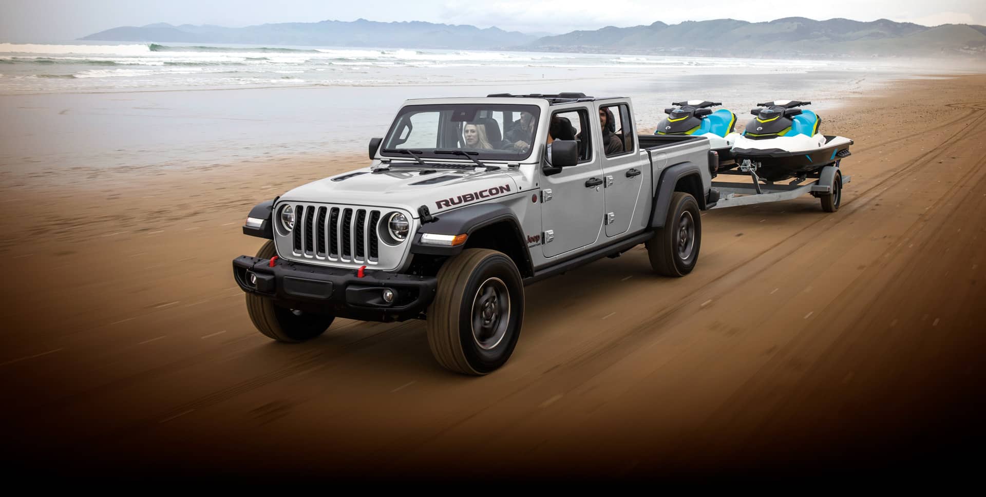 La Jeep Gladiator Rubicon 2022 remolcando dos motos de agua por una playa, cerca del mar.