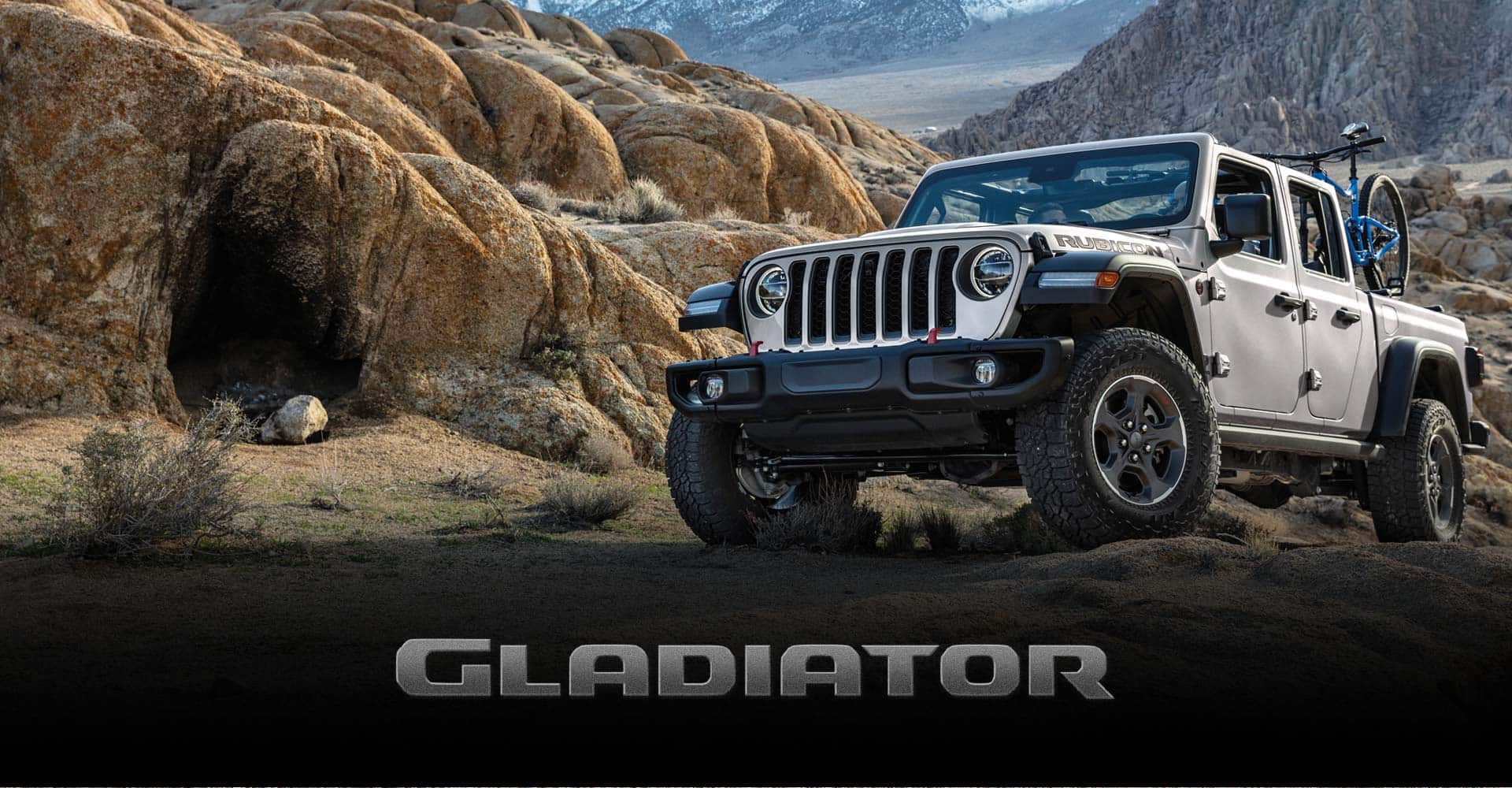Una vista frontal de tres cuartos de un Jeep Gladiator Rubicon 2022 gris con dos bicicletas de montaña en su caja, andando sobre rocas arenosas, con cadenas montañosas a la distancia. Gladiator.
