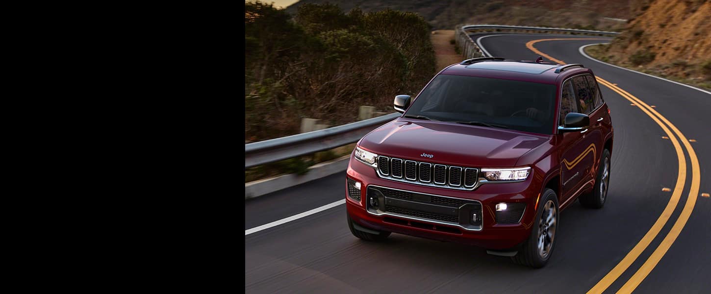 A 2022 Jeep Grand Cherokee with its headlamps on, taking a curve on a mountain road at dusk.