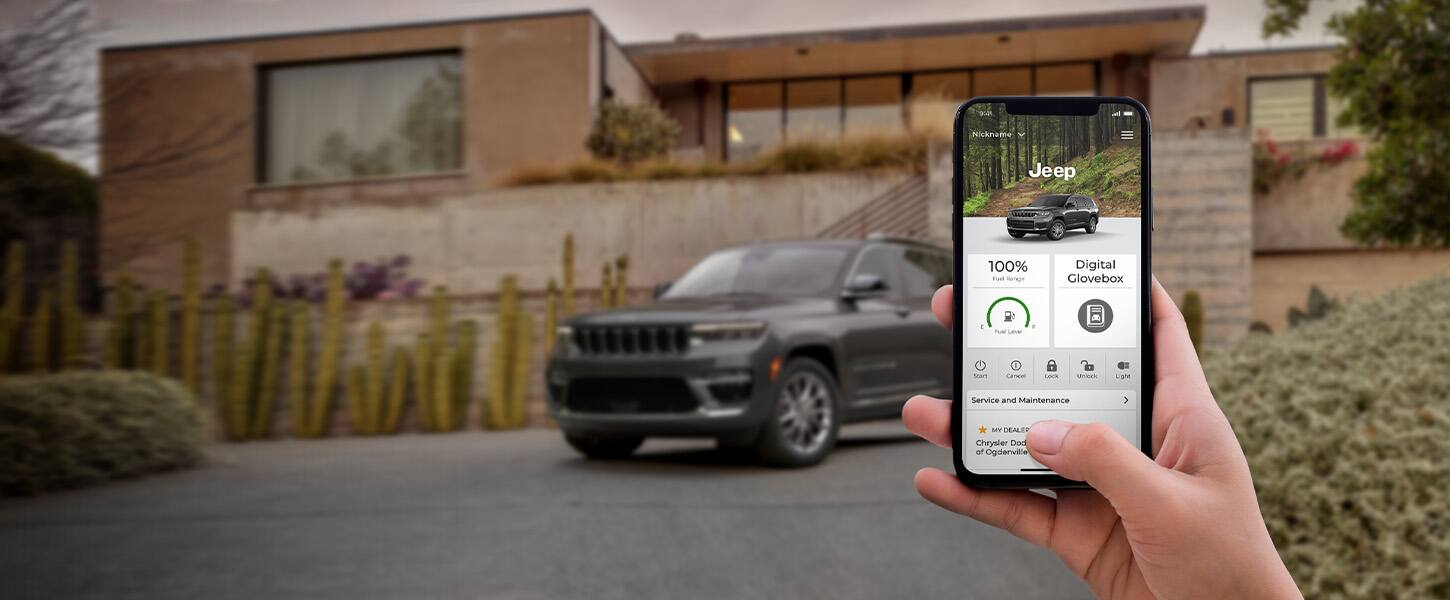 En primer plano, una mano que sostiene un smartphone que muestra la aplicación Jeep Connect. Al fondo, un Jeep Grand Cherokee L 2022.